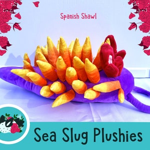 May include: Purple and yellow sea slug plush toy with a red flower on its head. The plush toy is labeled "Spanish Shawl" and "Sea Slug Plushies".