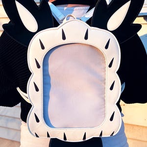 May include: A white and black backpack with a large clear window on the back. The backpack has a cartoonish design with large ears and a tail.