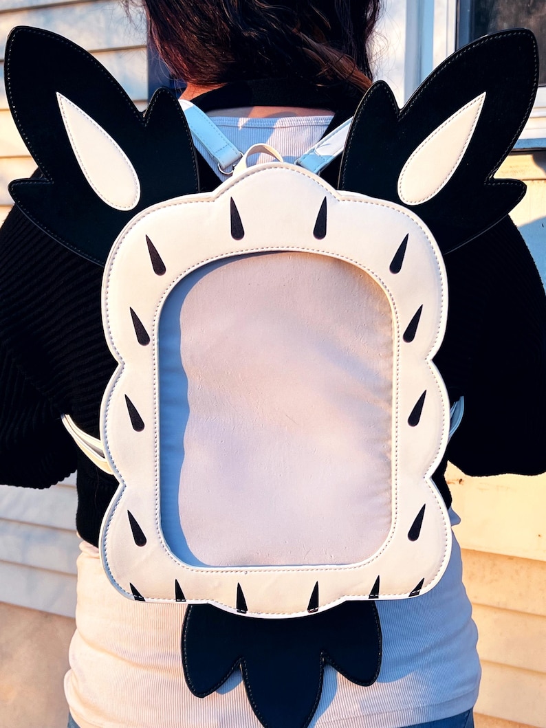 May include: A white and black backpack with a large clear window on the back. The backpack has a cartoonish design with large ears and a tail.