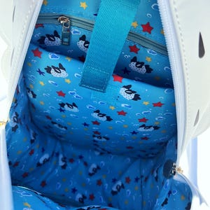 May include: The inside of a white pufferfish backpack with a blue lining. The lining has a pattern of pufferfish, stars, and bubbles. The backpack has a zippered pocket on the inside.