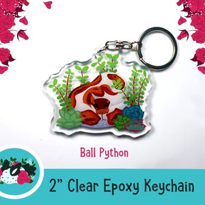 Może przedstawiać: Brelok z przezroczystego żywicy epoksydowej o wymiarach 5 cm przedstawiający węża boa dusiciela zwiniętego w łóżku z zielonych sukulentów. Brelok ma srebrny metalowy kółko do kluczy.