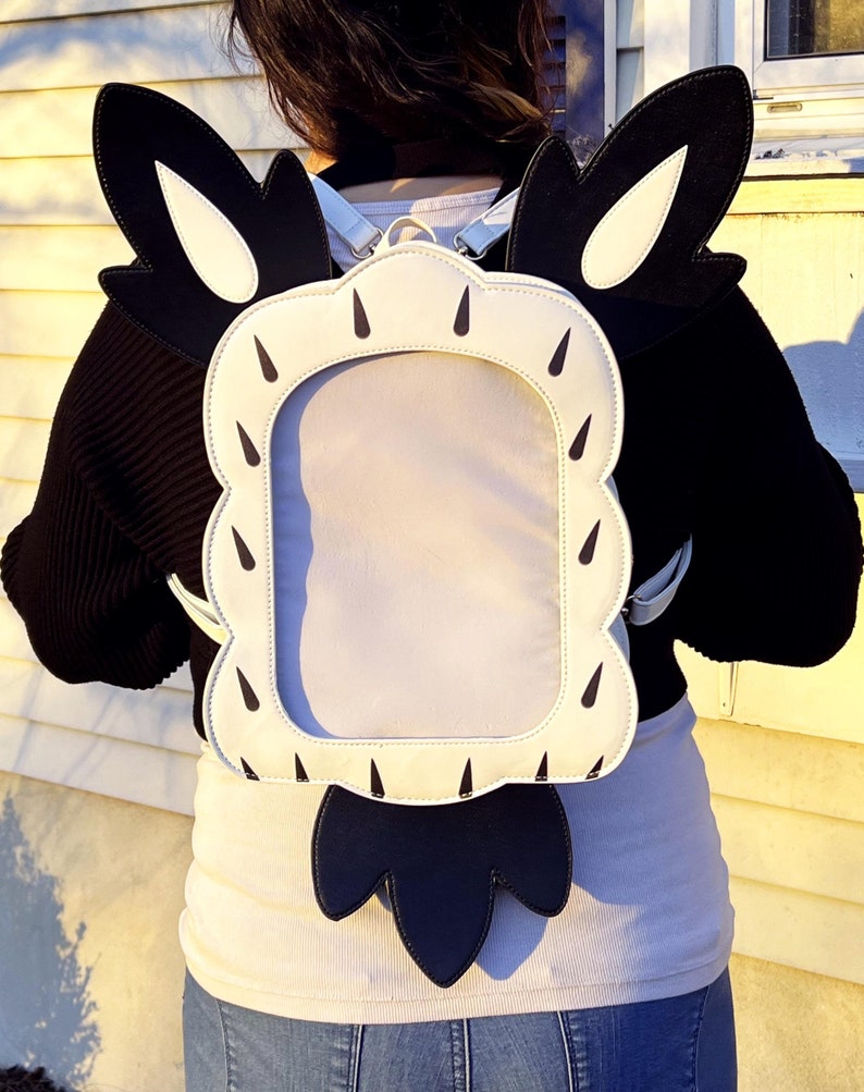 May include: A white and black backpack with a large rectangular window on the front. The backpack has black wings with white accents.