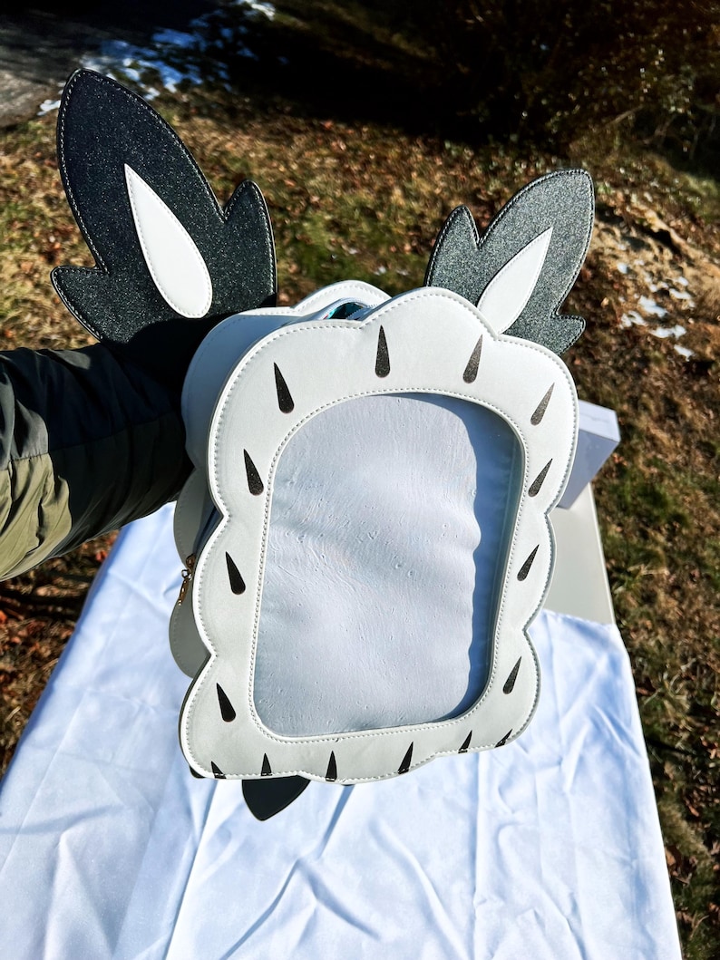 May include: White faux leather purse with black and white ears and black teardrop-shaped accents. The purse has a clear plastic window.