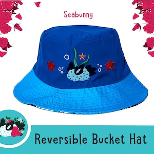 May include: A reversible bucket hat with a blue crown and a light blue brim. The hat features an embroidered design of a sea creature, starfish, and bubbles. The word "Seabunny" is above the hat.