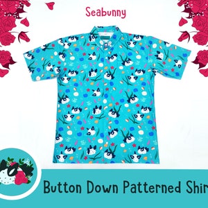 以下が含まれることがあります： 黒と白のウミウサギ、海藻、貝殻のパターンデザインが施されたターコイズブルーのボタンダウンシャツ。シャツは襟と半袖です。シャツの上に「Seabunny」というテキストが書かれており、シャツの下に「Button Down Patterned Shirt」というテキストが書かれています。