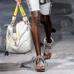 May include: A sleeveless blue vest with a layered lapel, a beige long-sleeve top, metallic silver shorts over patterned leggings, and a large white handbag with yellow handles. The ensemble includes dark sunglasses and grey lace-up sandals.