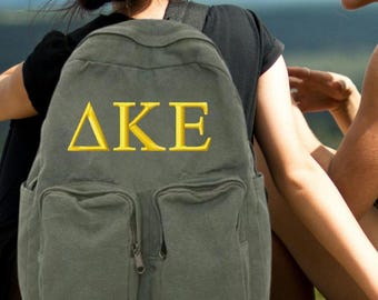 Peut inclure: Un sac à dos en toile gris foncé avec un grand motif brodé de lettres grecques jaunes qui lit "ΔΚΕ".