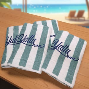 May include: Three white and teal striped beach towels with the text "Yall Yalla" in a script font. The towels are folded and stacked on a wooden surface, with a beach scene in the background.