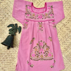 K&ouml;nnte beinhalten: Rosa besticktes Kleid mit kurzen &Auml;rmeln. Das Kleid hat Blumenstickereien in Rosa, Gr&uuml;n und Gold um den Halsausschnitt und am Oberteil. Auf der linken Seite des Kleides befindet sich eine dekorative Bronzeglocke.