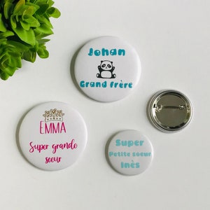 May include: Three round white button pins with colorful text and images. The top pin says "Johan Grand frère" with a black and white panda bear image. The middle pin says "Emma Super grande soeur" with a gold crown image. The bottom pin says "Super Petite soeur Inès" with a blue and white design.
