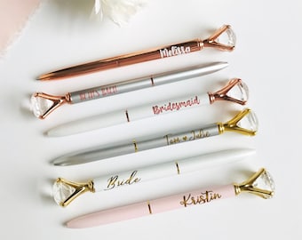 Puede incluir: Cinco bolígrafos personalizados con tapas en forma de diamante. Los bolígrafos son plateados, oro rosa y rosa con detalles dorados. Los bolígrafos están grabados con las palabras "Bride", "Maid of Honor", "Bridesmaid", "Tom & Julie" y "Kristin".