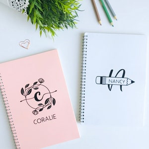 May include: Two spiral bound notebooks, one pink with a floral design and the name "CORALIE" and the other white with a pencil drawing and the name "NANCY".
