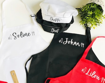 Könnte beinhalten: Drei personalisierte Schürzen in Weiß, Schwarz und Rot mit den Namen Soline, Johan und Lilou bestickt. Jede Schürze hat eine Tasche und ist mit einem kleinen Zweig Grün und einem Paar Kochutensilien verziert. Ein weißer Kochhut mit den Worten "Cheffe" und "Chef" bestickt ist ebenfalls enthalten.