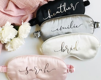 May include: Four satin sleep masks with different names written on them in gold and pink lettering. The names are "Heather", "Emilie", "Bride", and "Sarah".