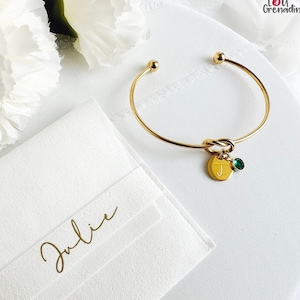 Peut inclure: Bracelet jonc en or avec un nœud et un pendentif en forme de lettre "J" et une pierre précieuse verte. Le bracelet est présenté dans une boîte blanche avec le nom "Julie" écrit en lettres dorées.