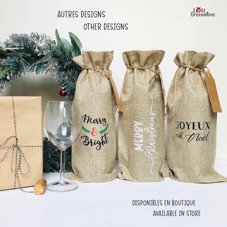 以下が含まれることがあります： クリスマスの刺繍が施された、麻製のワインボトル用ギフトバッグが3つ。バッグは白い表面に並べて置かれています。バッグには、「メリー＆ブライト」、「メリークリスマス」、「ジョワイユ・ノエル」の文字が刺繍されています。