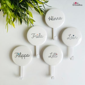 May include: Five white circular wall hooks with names in cursive script: Hanna, Frida, Leon, Filippa, and Lilli. Each hook has a small crown icon next to the name. Green leafy plant in the upper left corner.