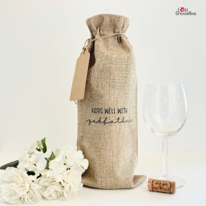 以下が含まれることがあります： 茶色の紙タグと「Pairs well with godfather」という文字が書かれた麻のワインボトルバッグ。