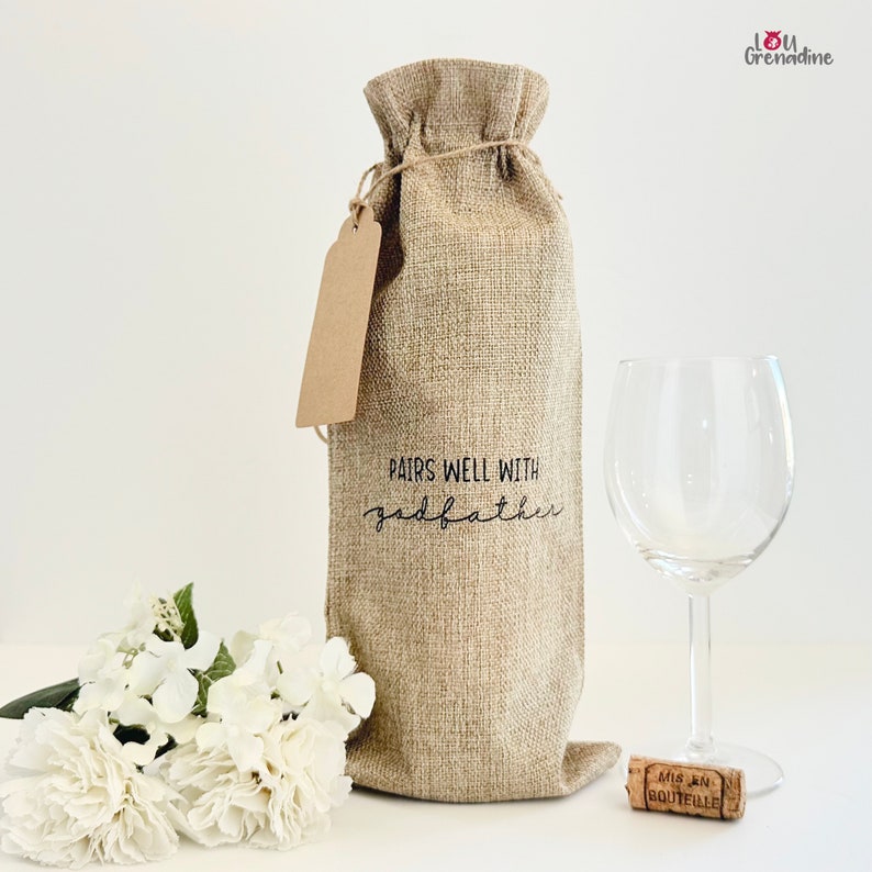 以下が含まれることがあります： 茶色の紙タグと「Pairs well with godfather」という文字が書かれた麻のワインボトルバッグ。