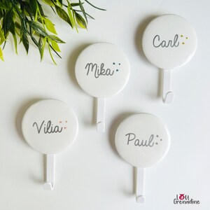 May include: Four white, round wall hooks with names: "Carl", "Mika", "Vilia", and "Paul" in gray script. Each hook has a small star design. The hooks are on a white surface with green leaves in the background.