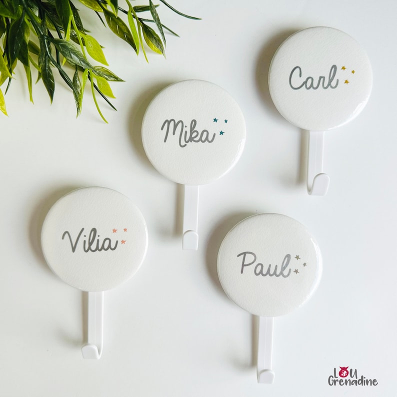 May include: Four white, round wall hooks with names: "Carl", "Mika", "Vilia", and "Paul" in gray script. Each hook has a small star design. The hooks are on a white surface with green leaves in the background.