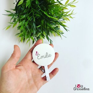 May include: A white, round compact mirror with the name "Emilie" in gray script and a pink butterfly graphic. The mirror has a white stand and is held in a hand, with green foliage in the background.