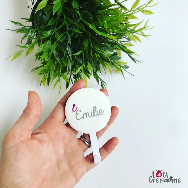 May include: A white, round compact mirror with the name "Emilie" in gray script and a pink butterfly graphic. The mirror has a white stand and is held in a hand, with green foliage in the background.
