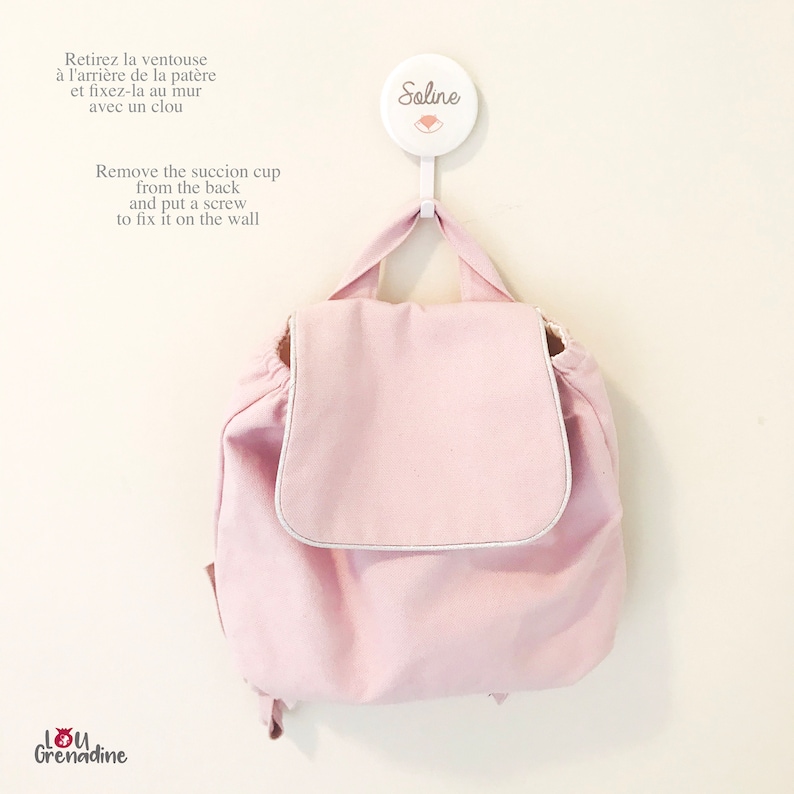 May include: A light pink backpack with a flap closure and silver trim hangs from a white hook. The hook has the name "Soline" printed on it. Text in French and English describes how to attach the hook to the wall.