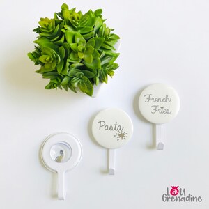 May include: White wall hooks with the words "Pasta" and "French Fries" printed on them. The hooks have a circular design and a small hook at the bottom. A small green plant in a white pot is in the background.