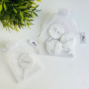 May include: White personalized round favors with names in silver script, packaged in sheer white bags. The favors have a handle and are displayed on a white surface. The image includes a tag with the text "I love Grenadine".