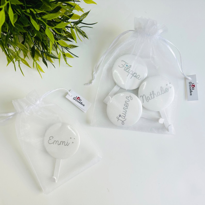 May include: White personalized round favors with names in silver script, packaged in sheer white bags. The favors have a handle and are displayed on a white surface. The image includes a tag with the text "I love Grenadine".
