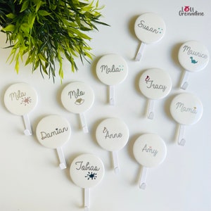 May include: White round wall hooks with names like Susan, Mila, and Tobias printed in gray. Each hook has a small decorative element, such as a dinosaur or paint splatter. The hooks are arranged on a white surface with a green plant in the background.