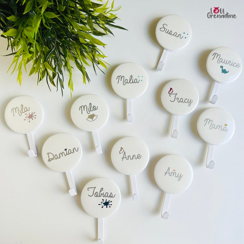 May include: White round wall hooks with names like Susan, Mila, and Tobias printed in gray. Each hook has a small decorative element, such as a dinosaur or paint splatter. The hooks are arranged on a white surface with a green plant in the background.