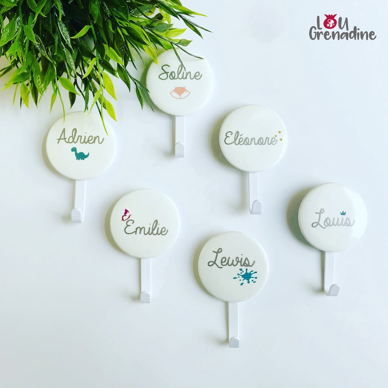 May include: White, round wall hooks with names and small illustrations. The hooks are labeled with the names: Adrien, Soline, &Eacute;l&eacute;onore, Emilie, Lewis, and Louis. Each hook has a different design, including a dinosaur, fox, stars, butterfly, paint splatter, and crown.
