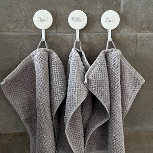 May include: Three gray washcloths hang from white hooks on a tiled wall. Each hook has a name printed on it: No&eacute;, Milla, and Luca. The washcloths are folded and have a textured pattern.