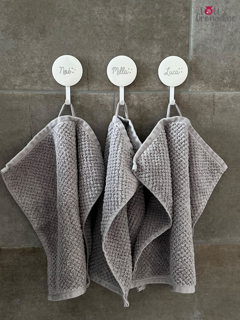 May include: Three gray washcloths hang from white hooks on a tiled wall. Each hook has a name printed on it: No&eacute;, Milla, and Luca. The washcloths are folded and have a textured pattern.