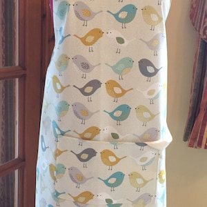 May include: A cream-coloured apron featuring a repeating pattern of colourful birds in blue, yellow, grey, and green. The apron has a large front pocket and white straps. The apron is displayed on a mannequin.