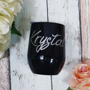 Puede incluir: Un vaso de vino de acero inoxidable negro con el nombre "Krystal" grabado en letras plateadas.