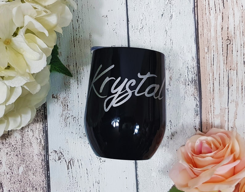 Puede incluir: Un vaso de vino de acero inoxidable negro con el nombre "Krystal" grabado en letras plateadas.
