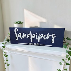 May include: A rectangular navy blue wooden sign featuring the white text Sandpipers in a cursive script font, with the text EST 2025 printed in a sans-serif font below, underlined by a horizontal white line spanning the width of the sign.