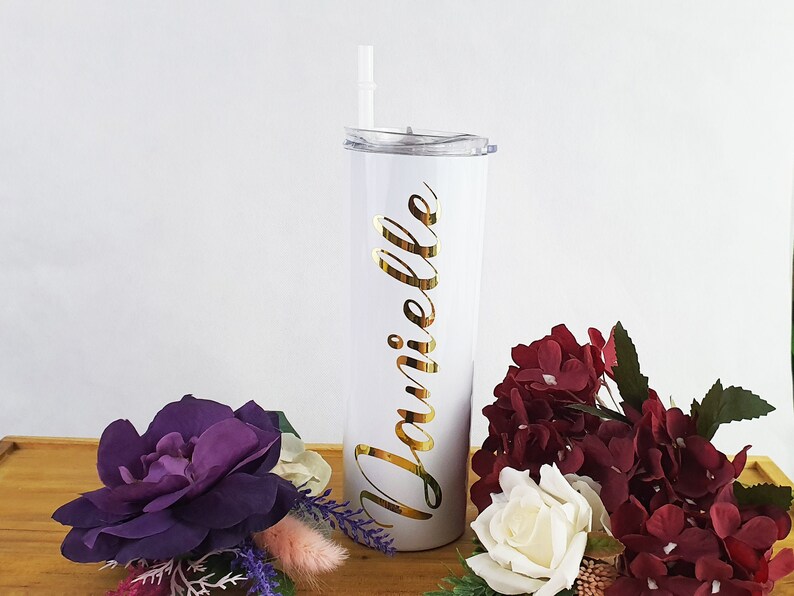 K&ouml;nnte beinhalten: Ein wei&szlig;er Edelstahlbecher mit einer goldenen Aufschrift "Danielle". Der Becher hat einen durchsichtigen Deckel und einen Strohhalm. Der Becher steht auf einer Holzoberfl&auml;che mit Blumen.