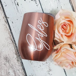 Puede incluir: Un vaso de vino de acero inoxidable color oro rosa con el nombre "Giles" grabado en letras blancas.