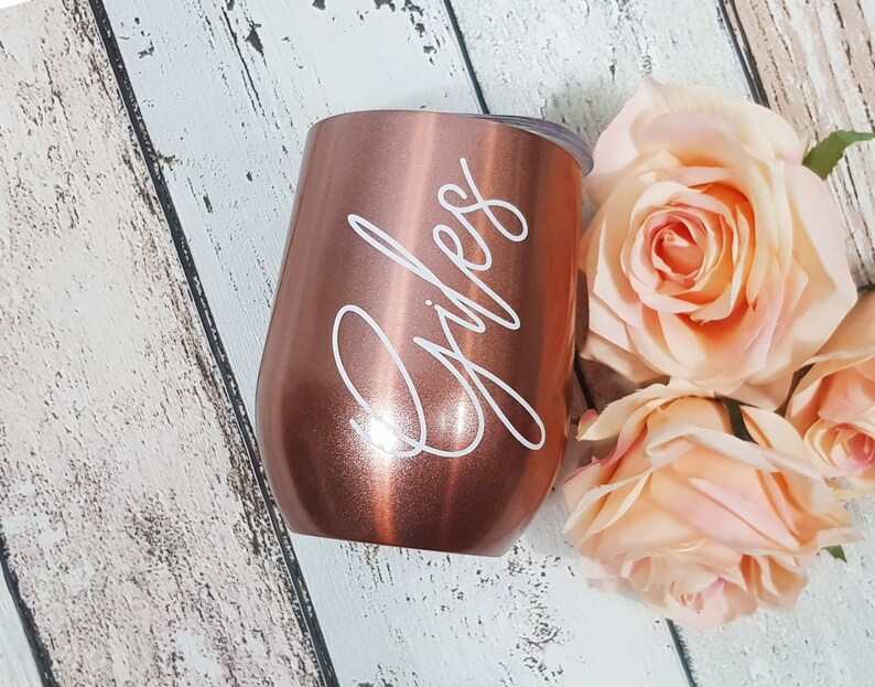 Puede incluir: Un vaso de vino de acero inoxidable color oro rosa con el nombre "Giles" grabado en letras blancas.