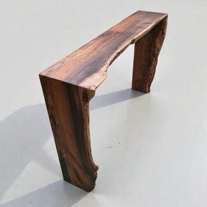 May include: A handcrafted console table made from a single piece of dark brown wood. The table has a live edge on the top and sides, with a natural, organic shape. The table is approximately 152 cm long and 38 cm wide.