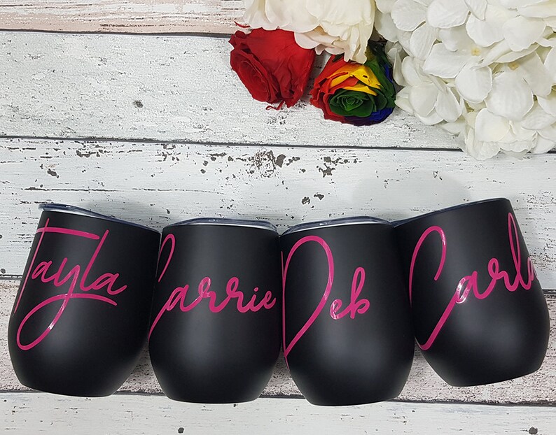 Puede incluir: Cuatro vasos de vino de acero inoxidable negro con letras rosas. Los vasos est&aacute;n personalizados con los nombres Tayla, Carrie, Deb y Carla.