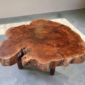 May include: A low coffee table featuring a natural live edge wood slab top with a warm brown finish, visible wood grain, and organic irregular edges. The table is supported by three short, cylindrical wooden legs attached to the underside.