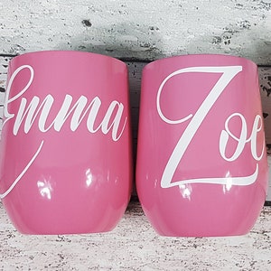 Puede incluir: Dos vasos de vino de acero inoxidable rosa con letras de vinilo blanco. Los vasos est&aacute;n personalizados con los nombres "Emma" y "Zoe".
