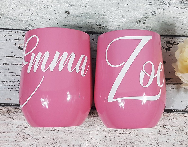 Puede incluir: Dos vasos de vino de acero inoxidable rosa con letras de vinilo blanco. Los vasos est&aacute;n personalizados con los nombres "Emma" y "Zoe".