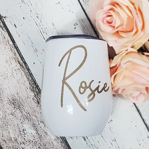 Puede incluir: Un vaso de vino de acero inoxidable blanco con el nombre "Rosie" en purpurina dorada en la parte delantera.