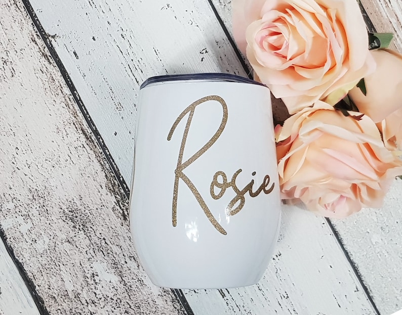 Puede incluir: Un vaso de vino de acero inoxidable blanco con el nombre "Rosie" en purpurina dorada en la parte delantera.